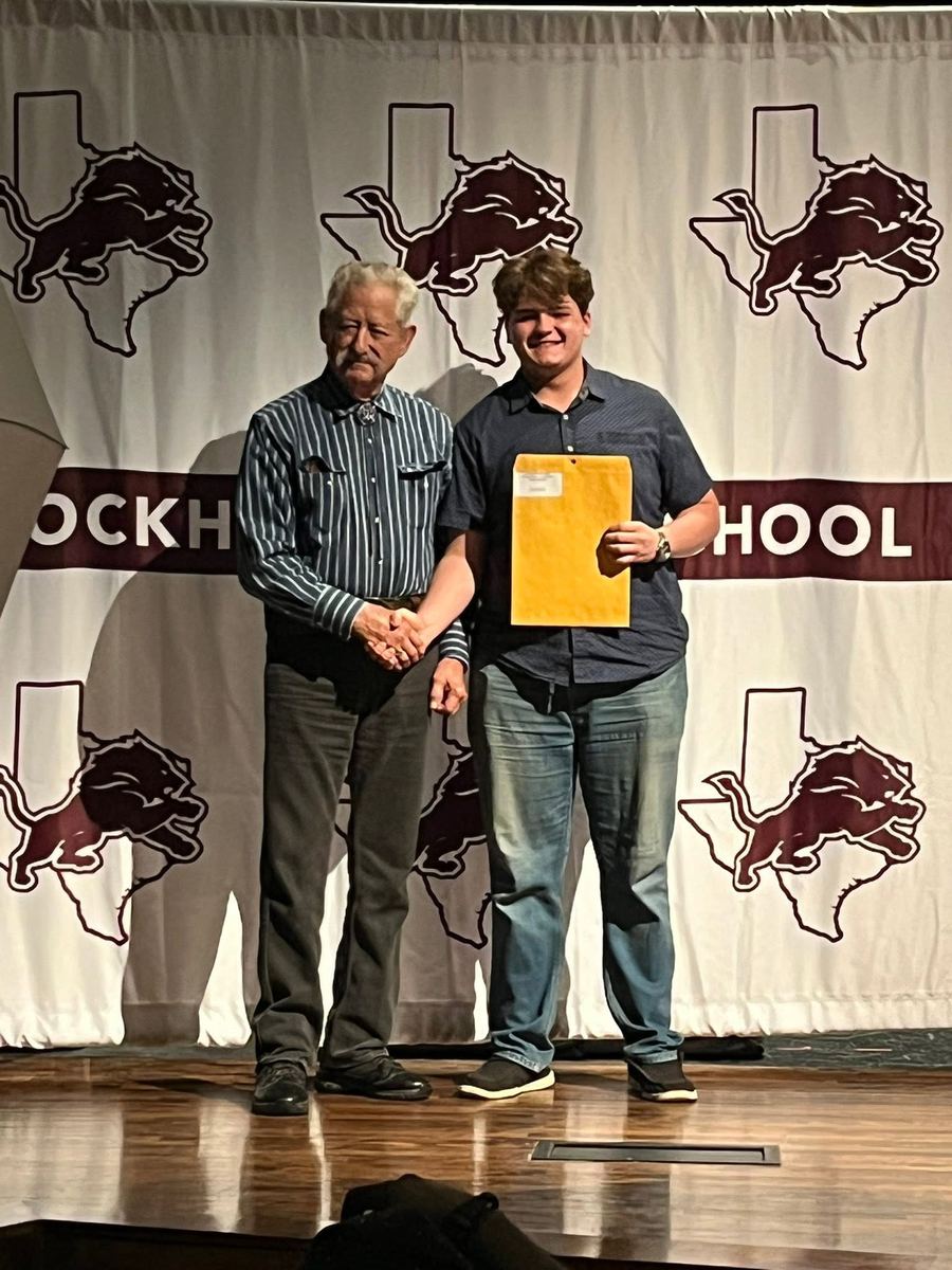 A lodge member shakes hands with a student holding an envelope on stage at Lockhart High School